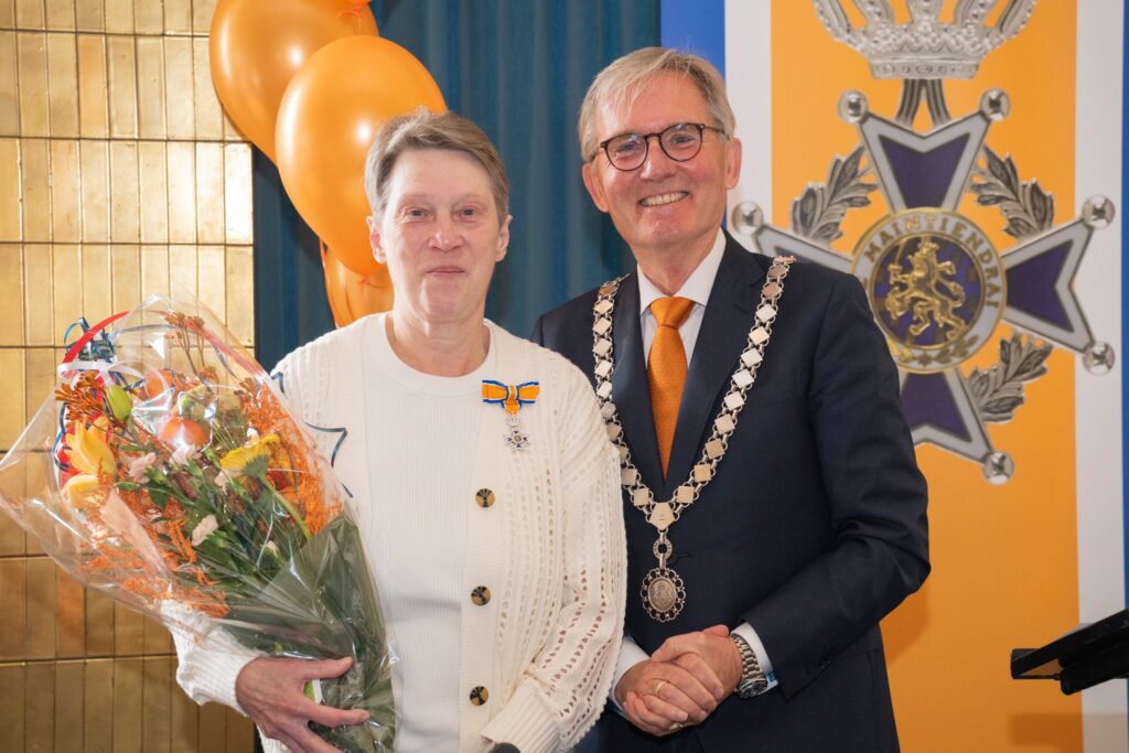 vrouw met koninklijke onderscheiding op de borst en bos bloemen op de arm staat glimlachend naast bebrilde burgemeester van Hilversum nadat zij is onderscheiden voor haar diensten als vrijwilliger